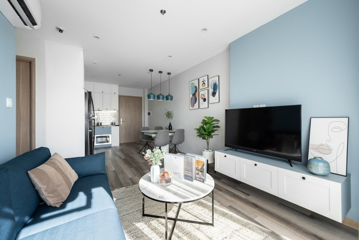 Phòng khách đón ánh sáng tuyệt đẹp từ logia căn hộ, thiết kế vuông vức với một sofa dài, bàn tròn, kệ tivi đơn giản và hiện đại.